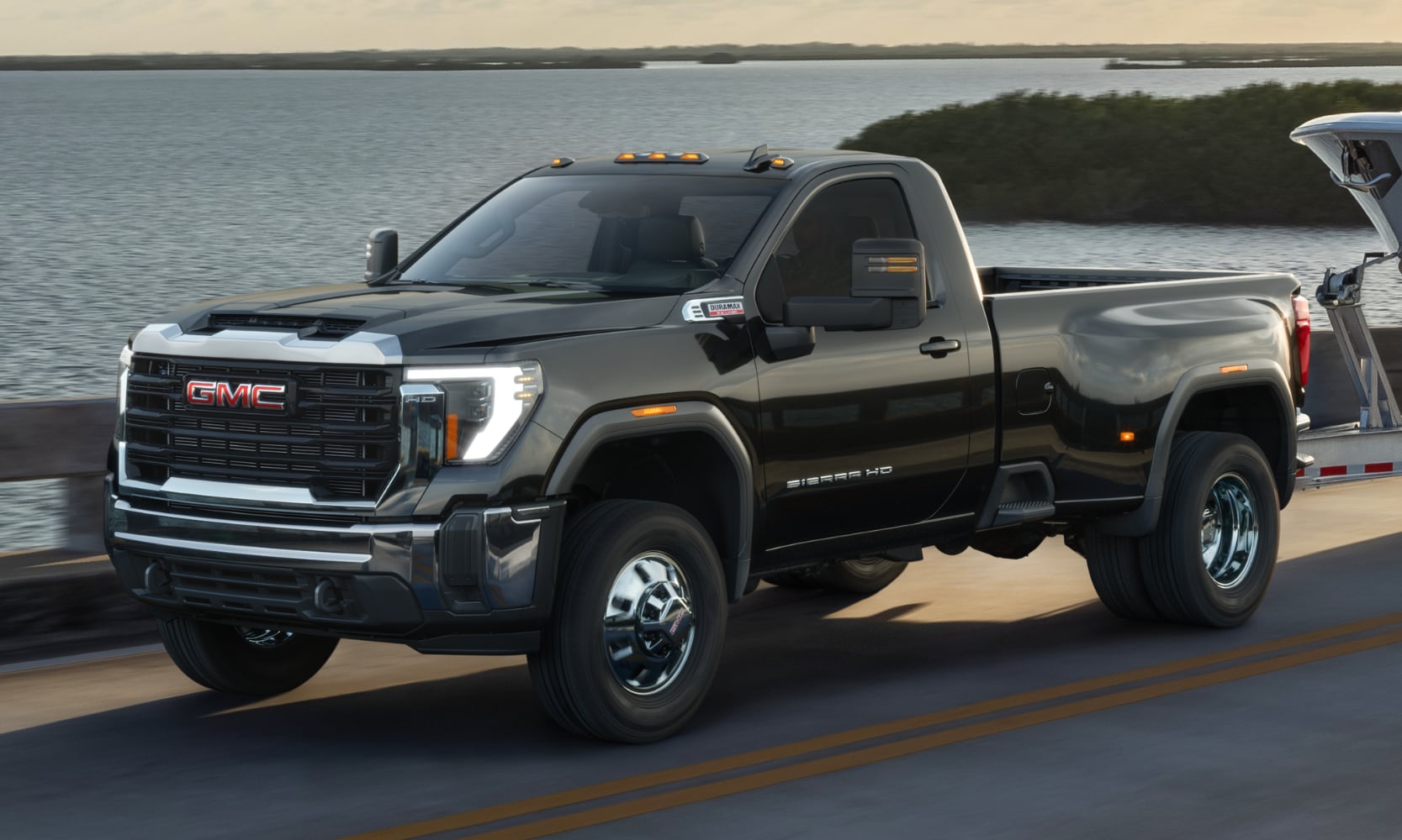 Closeup image of a black 2026 GMC Sierra HD Pro towing a boat on a waterside freeway