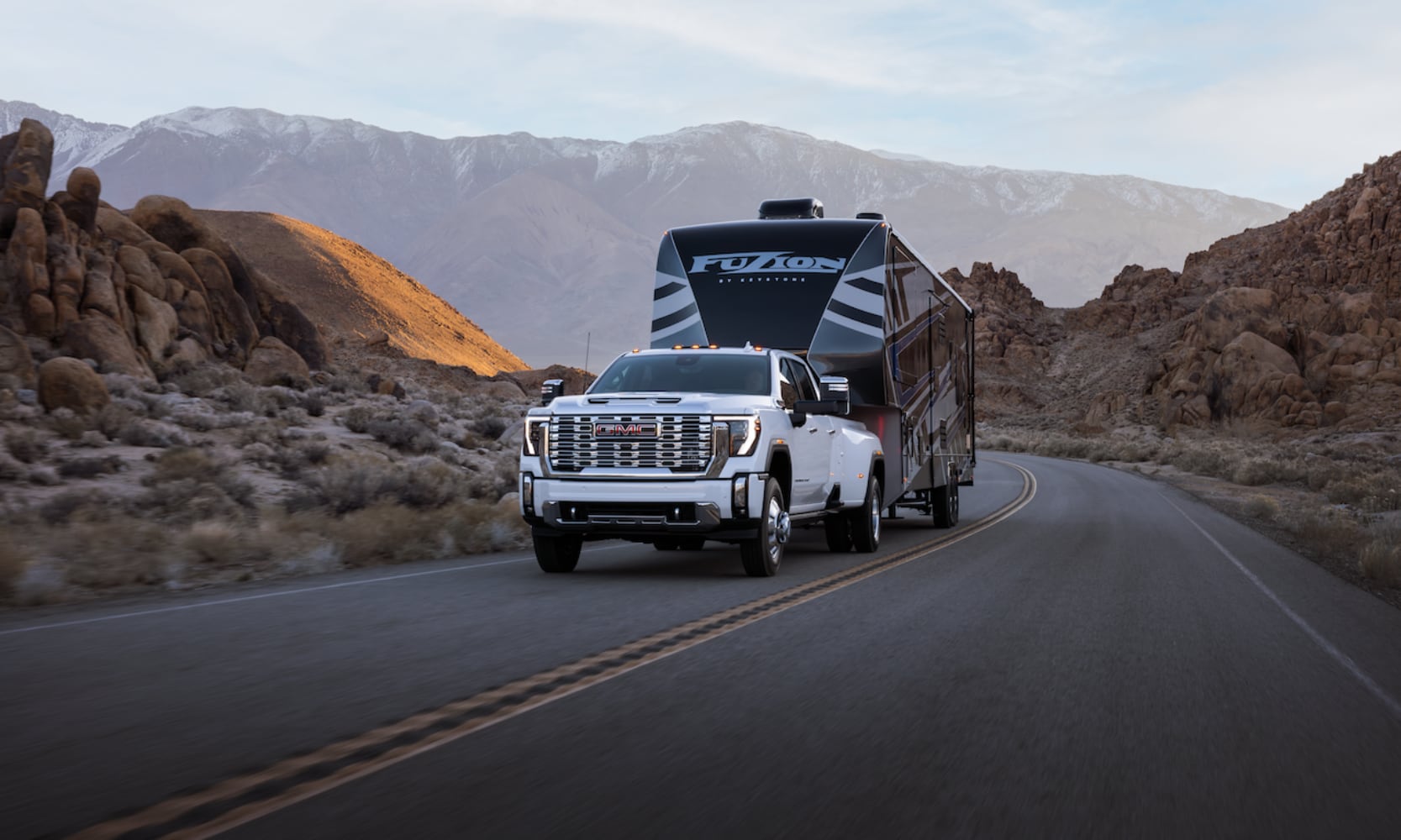 Exterior front view image of a white 2026 GMC Sierra 3500 HD Denali towing a camping trailer on a mountain freeway