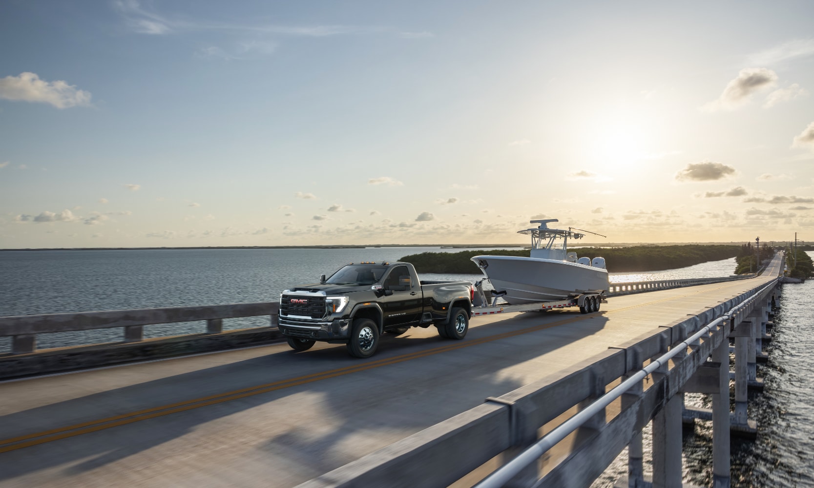 Exterior far-view mage of a black 2026 GMC Sierra 3500 HD Pro towing a small boat on a waterfront freeway