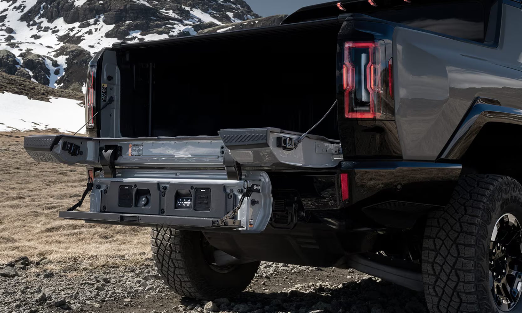 Closeup exterior image of the 2026 GMC Hummer EV Pickup opened MultiPro Tailgate with a snowy mountain in the background