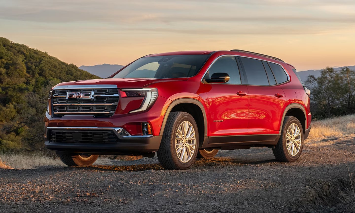 Exterior front-view image of a red 2026 GMC Acadia parked in a grassy mountain setting