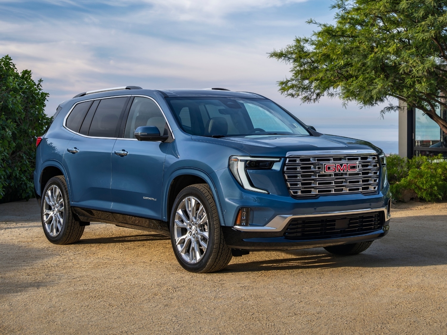 a blue 2025 GMC Acadia Denali SUV parked outside a sleek contemporary home overlooking a cliff