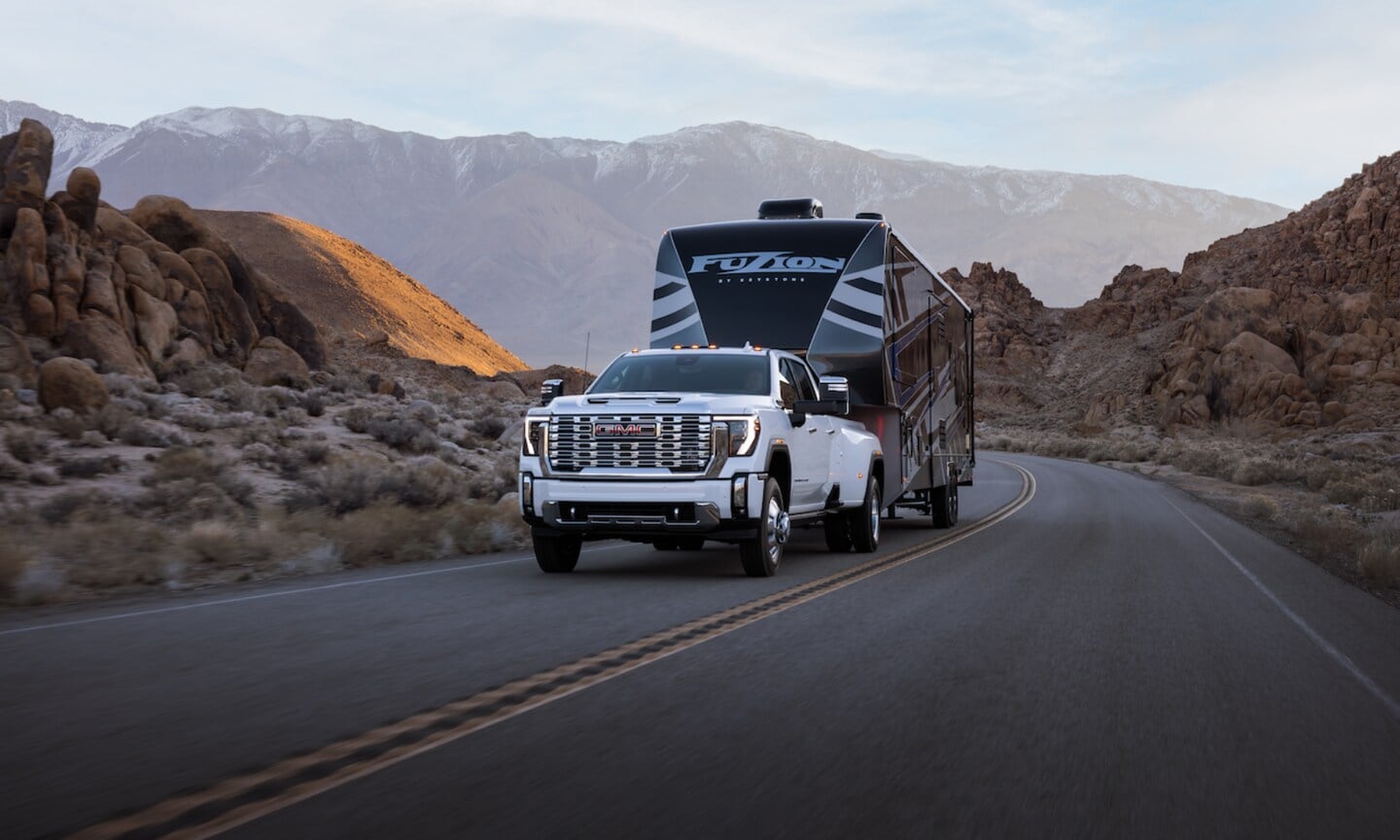 Exterior front-view image of a white 2026 GMC Sierra HD 3500 towing a camping trailer on a mountainous freeway