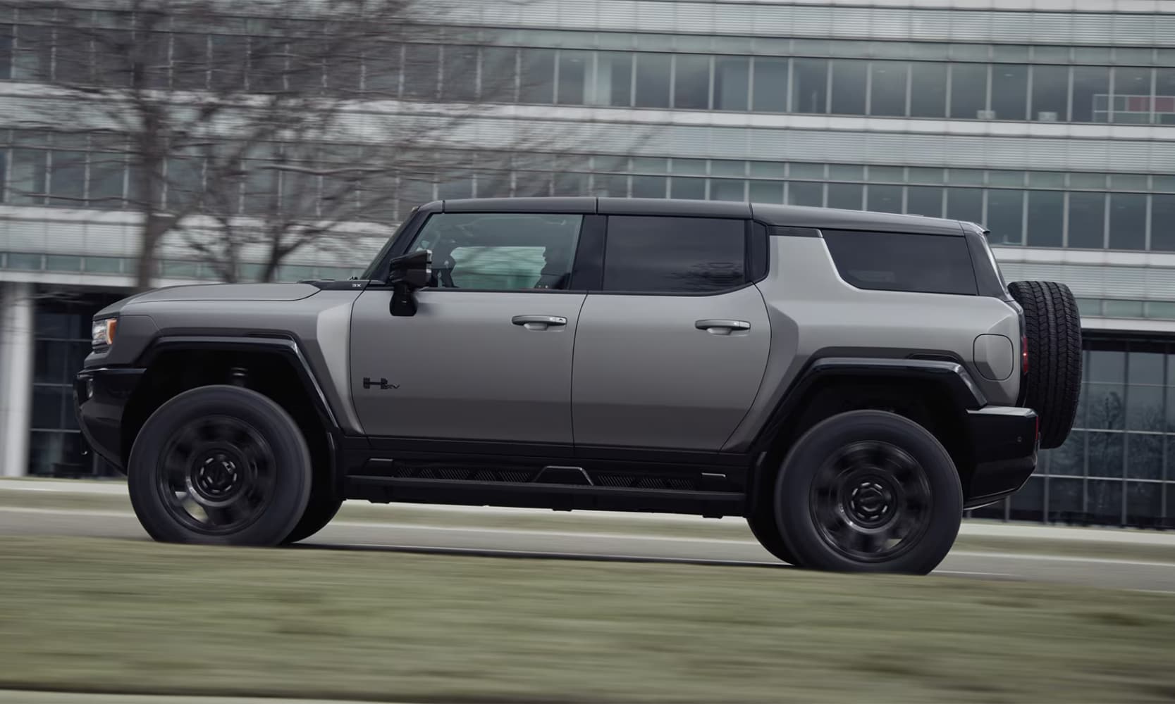 Exterior side image of a light gray 2026 GMC Hummer EV SUV driving past a commercial building