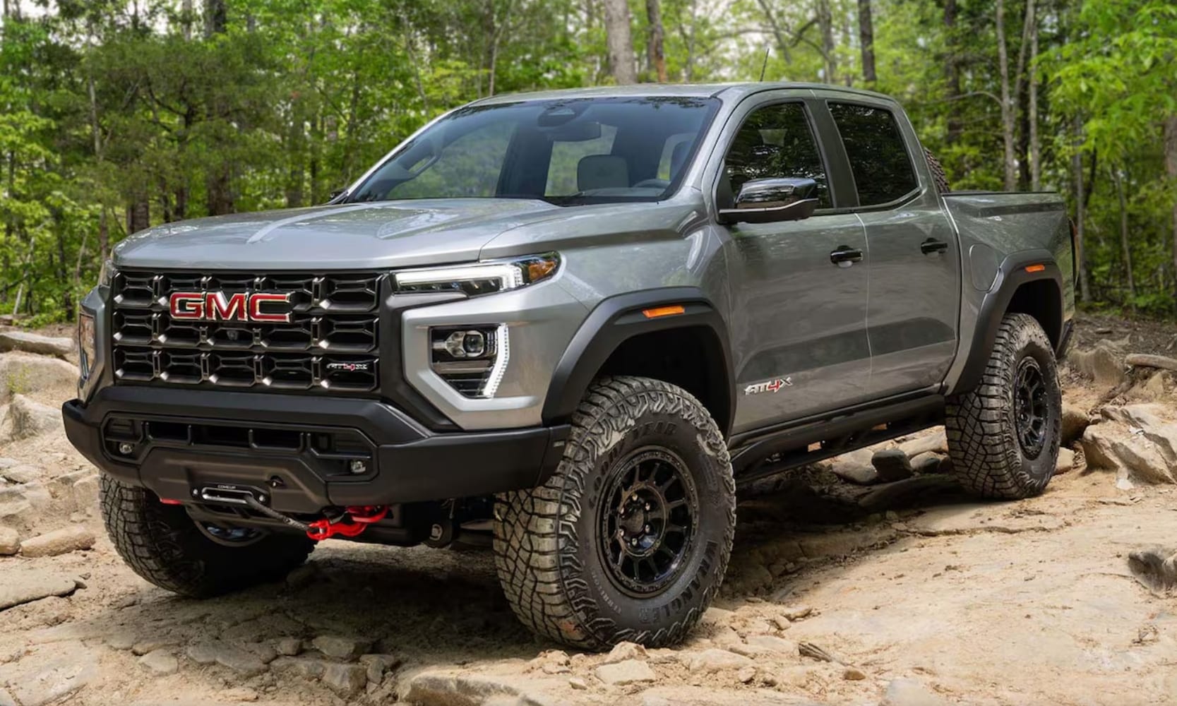 Exterior image of a light gray 2024 GMC Canyon AT4X AEV Edition parked in a forest landscape during the day