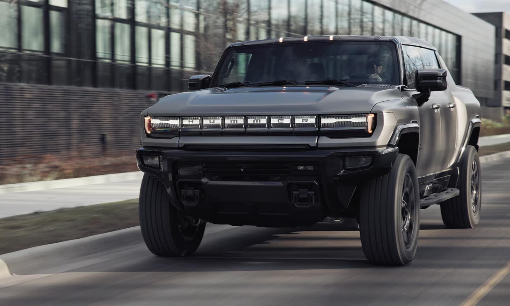 Exterior front image of a gray 2026 GMC Hummer EV Pickup driving down a road passing a commercial building