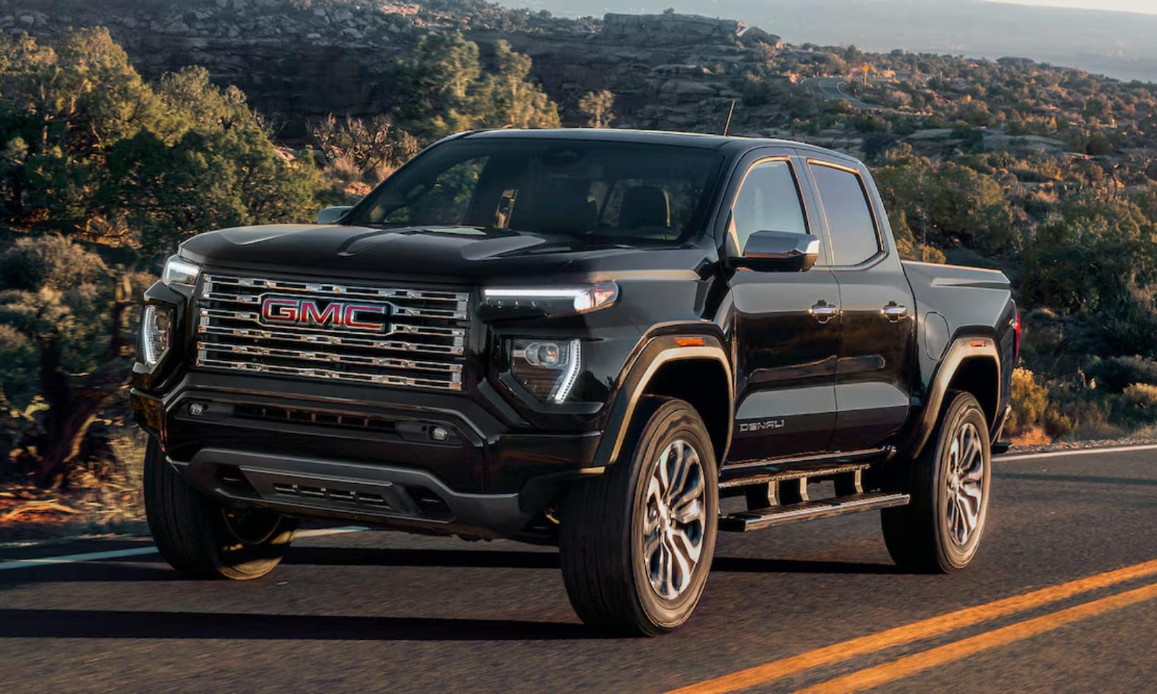 Exterior image of a black 2026 GMC Canyon Denali driving on an open tree-lined freeway