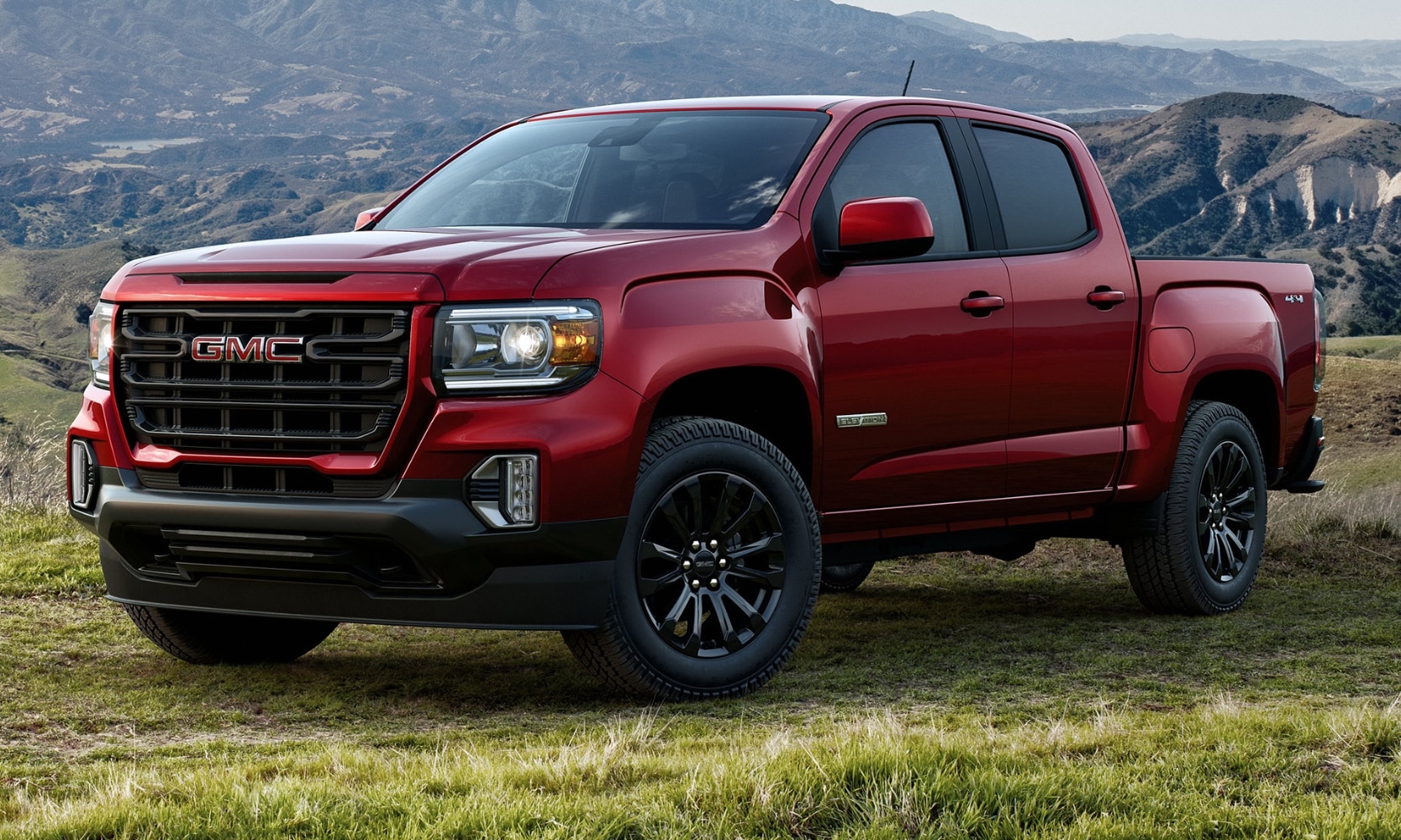Exterior side angle image of a red 2022 GMC Canyon Elevation parked on a grassy hill with mountains in the background