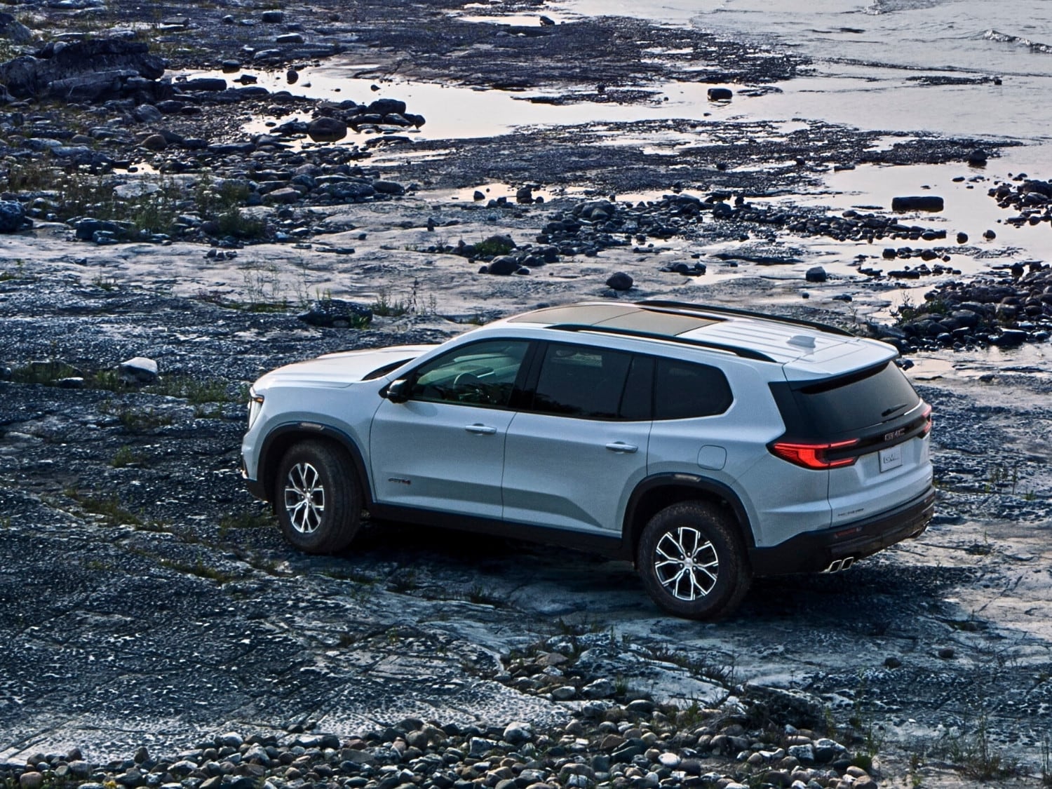 a serene aerial view of a white 2025 GMC Acadia SUV parked on a rocky shore by a beautiful lake