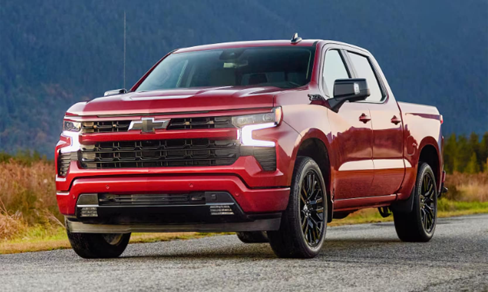 Exterior front image of a red 2025 Chevy Silverado RST parked on a road in a mountain landscape with its lights on