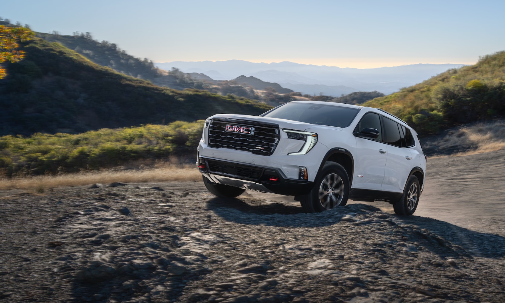Exterior front angle image of a white 2022 GMC Acadia AT4 driving up a steep rocky incline in a mountainous setting