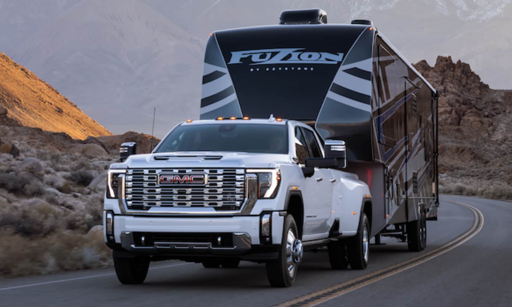 Closeup exterior image of a white 2026 GMC Sierra HD 3500 towing a camping trailer on an open mountainous freeway