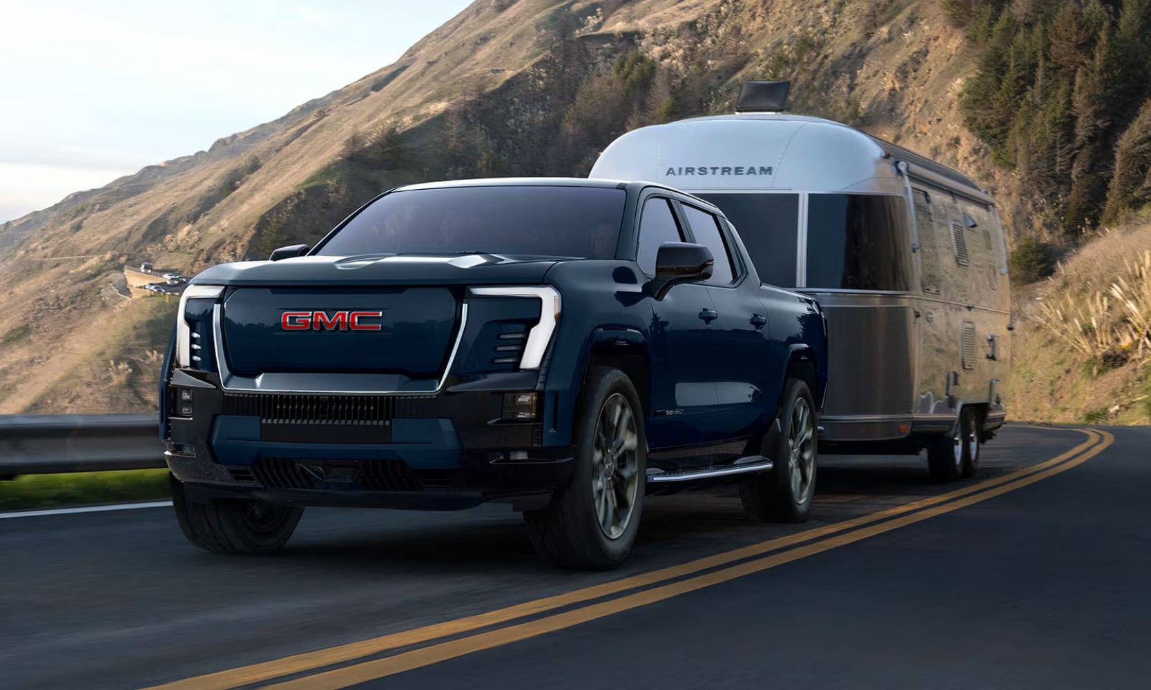 Exterior front image of a dark blue 2026 GMC Sierra EV Denali towing a camping trailer on a mountainous freeway