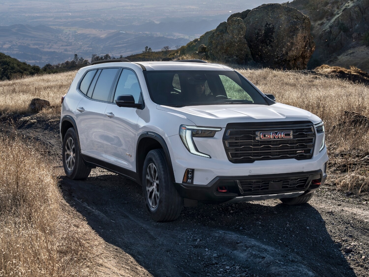 a white 2025 GMC Acadia AT4 SUV driving through the mountains on a dirt road