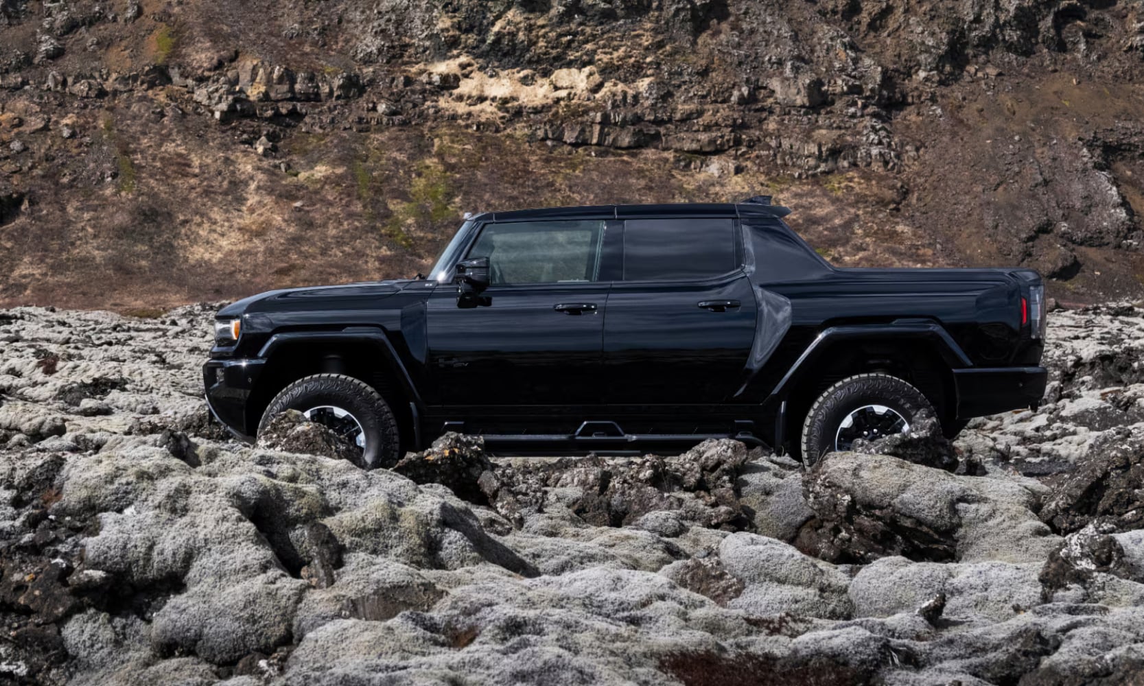 Side exterior image of a black 2025 GMC Hummer EV Truck off-roading through large boulders in a mountainous landscape