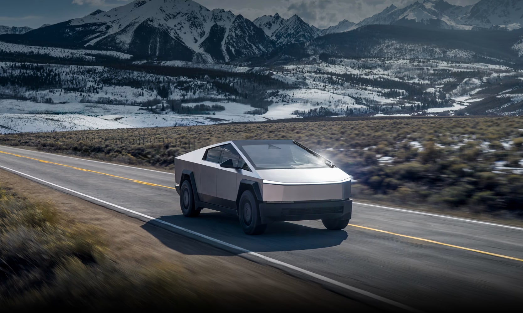 Exterior front angle image of a silver 2025 Tesla Cybertruck driving on an open freeway in a snowy mountain landscape
