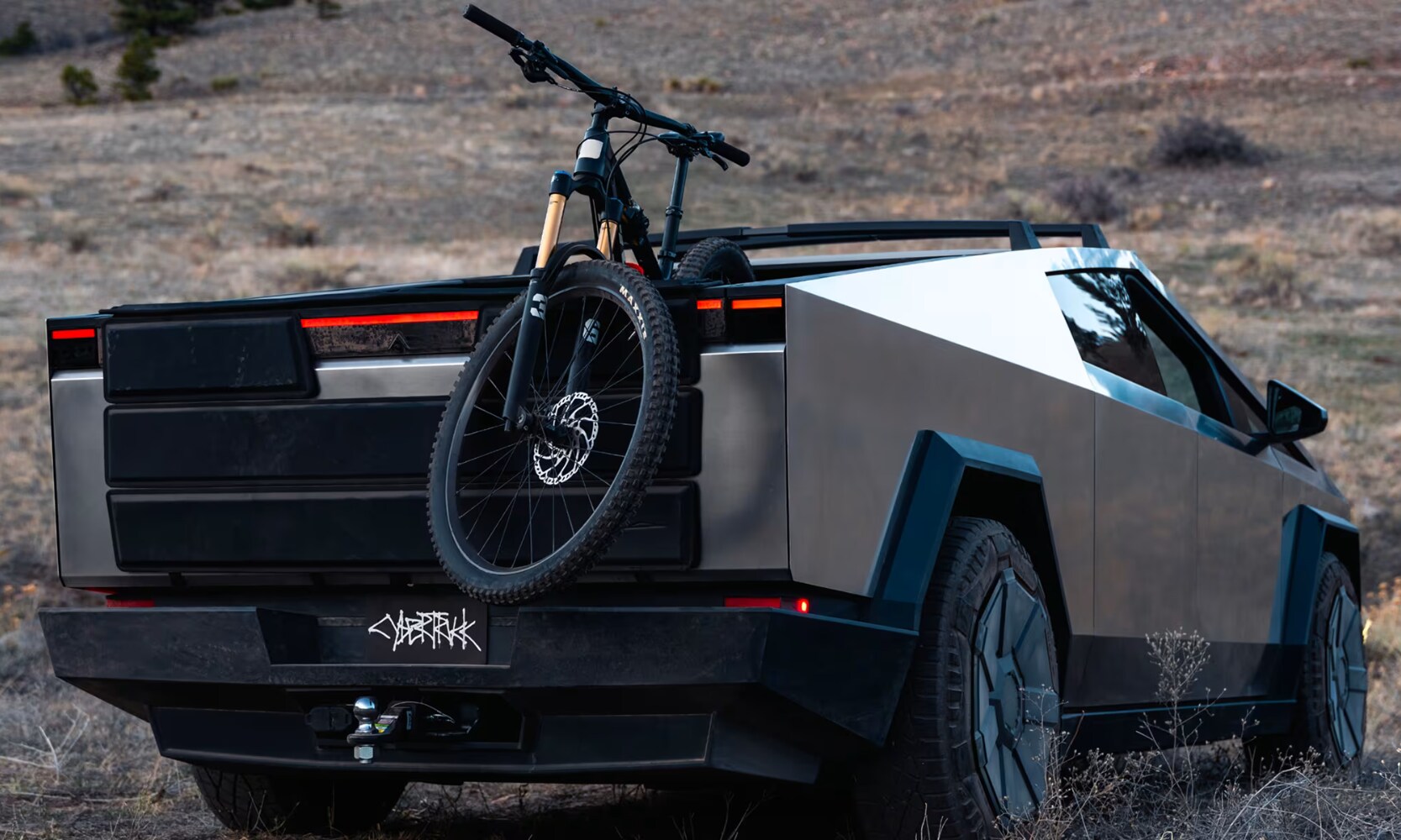 Rear-view image of a silver 2025 Tesla Cybertruck driving through a grass field with a bicycle in its trunk
