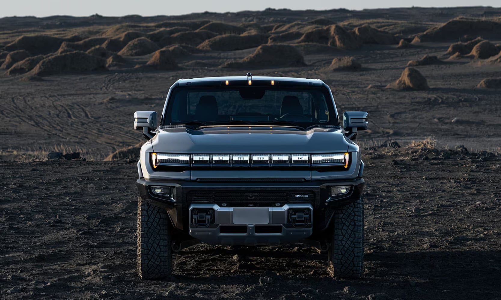 Front view image of a dark gray 2025 GMC Hummer EV Truck parked in a dirt hill landscape