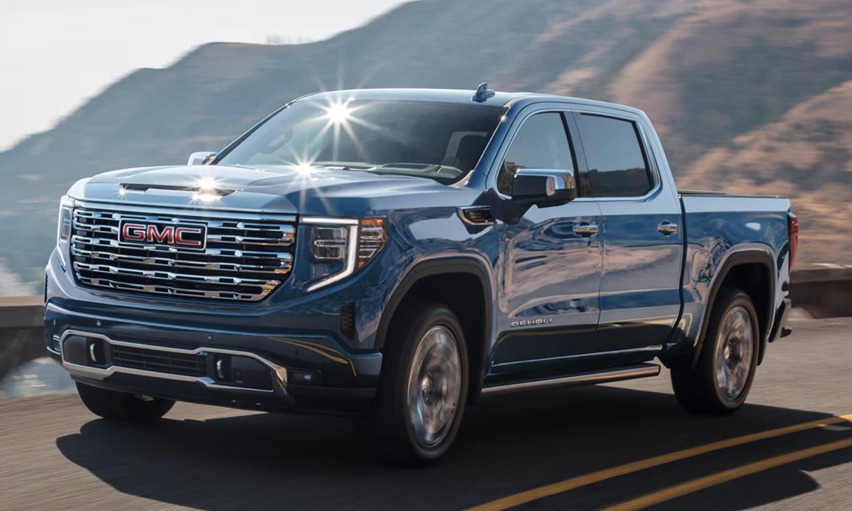 Closeup exterior image of a blue 2026 GMC Sierra 1500 Denali driving on a mountainside freeway