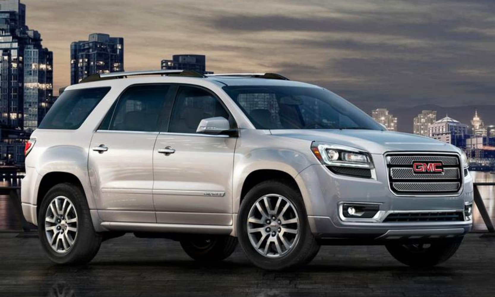 Exterior side image of a white 2015 GMC Acadia Denali parked in a cityscape setting at night