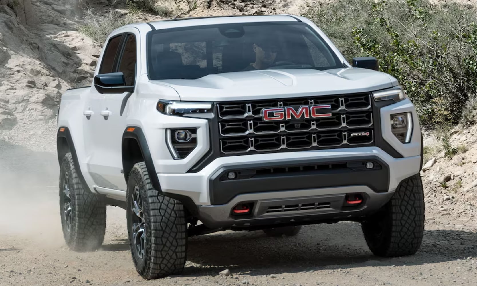Exterior front image of a white 2024 GMC Canyon AT4 driving up a dirt hill with dirt kicking up behind the rear tires