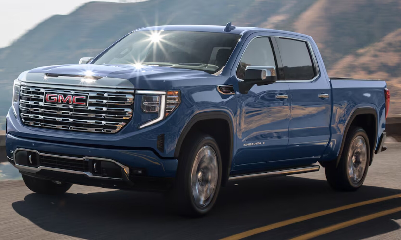 Exterior image of a blue 2025 GMC Sierra 1500 Denali driving on a freeway with mountains in the background