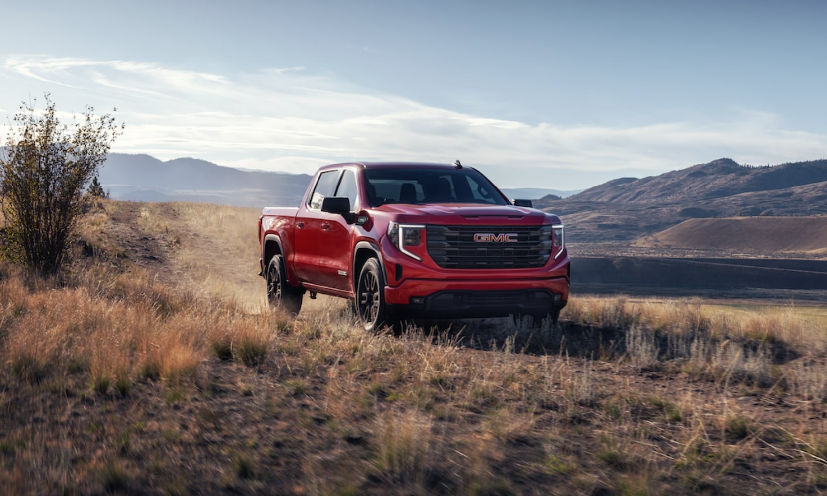 Exterior front image of a red 2025 GMC Sierra 1500 Elevation driving through a grass field in a mountain landscape