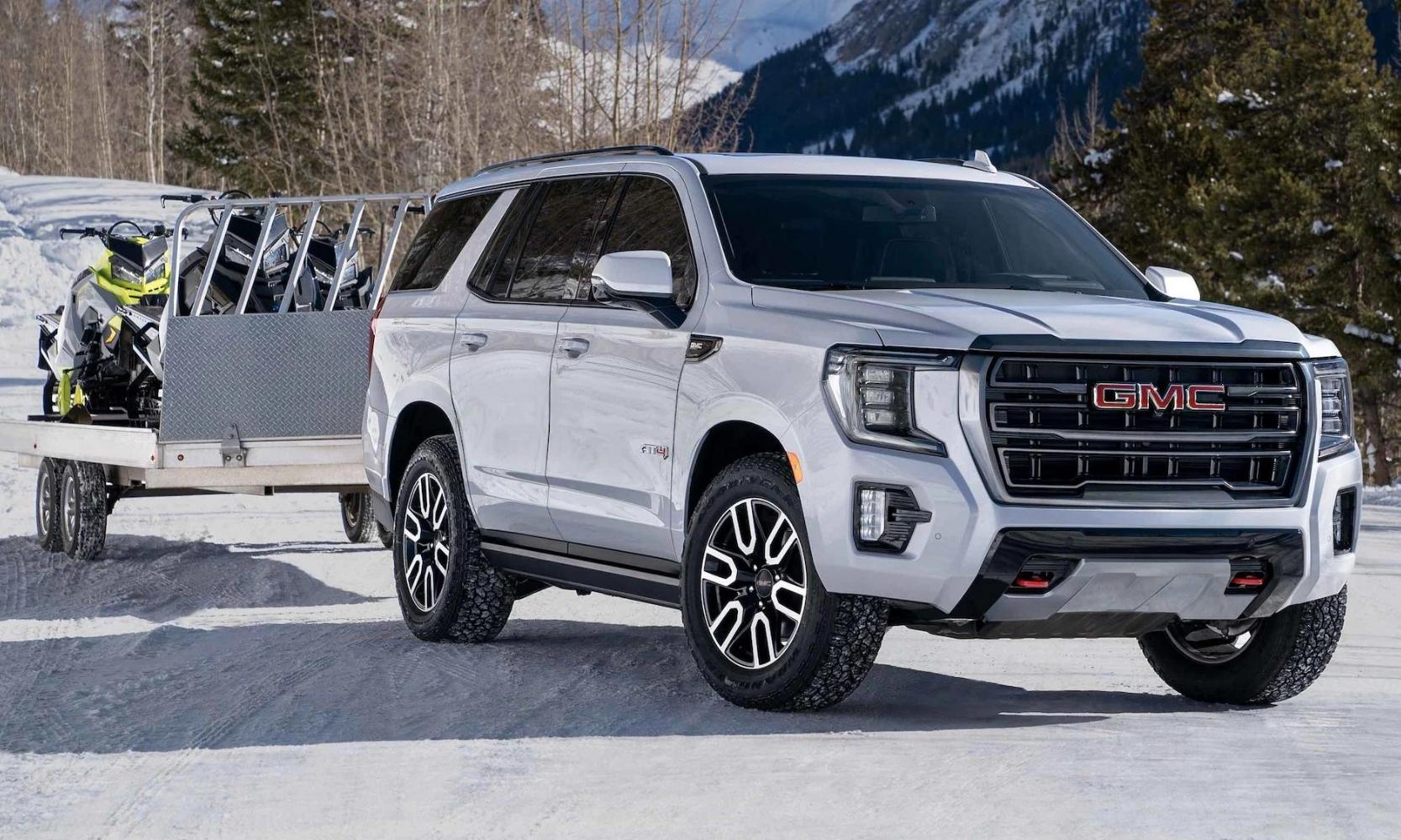 a white 2023 GMC Yukon AT4 towing a trailer loaded with 4 snowmobiles on a snowy mountain