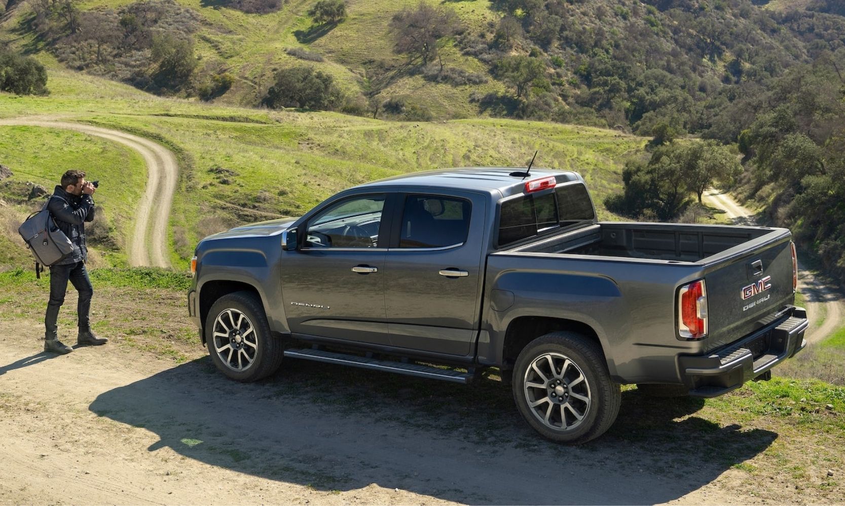 Aerial exterior view of a parked gray 2022 GMC Canyon Denali truck as the driver stands behind it taking photos