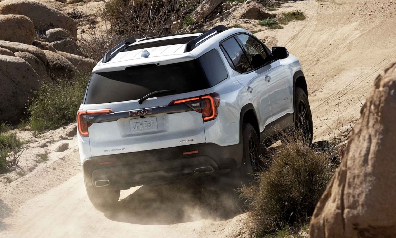 Rear view of a white 2022 GMC Acadia AT4 showing off its efficient engine performance as it drives uphill on a dirt road slope
