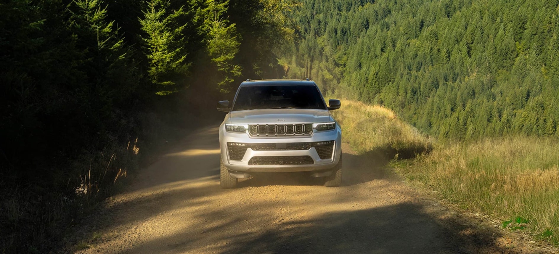 A front view of a silver Jeep Grand Cherokee driving over a dirt road on a hill in a forest.