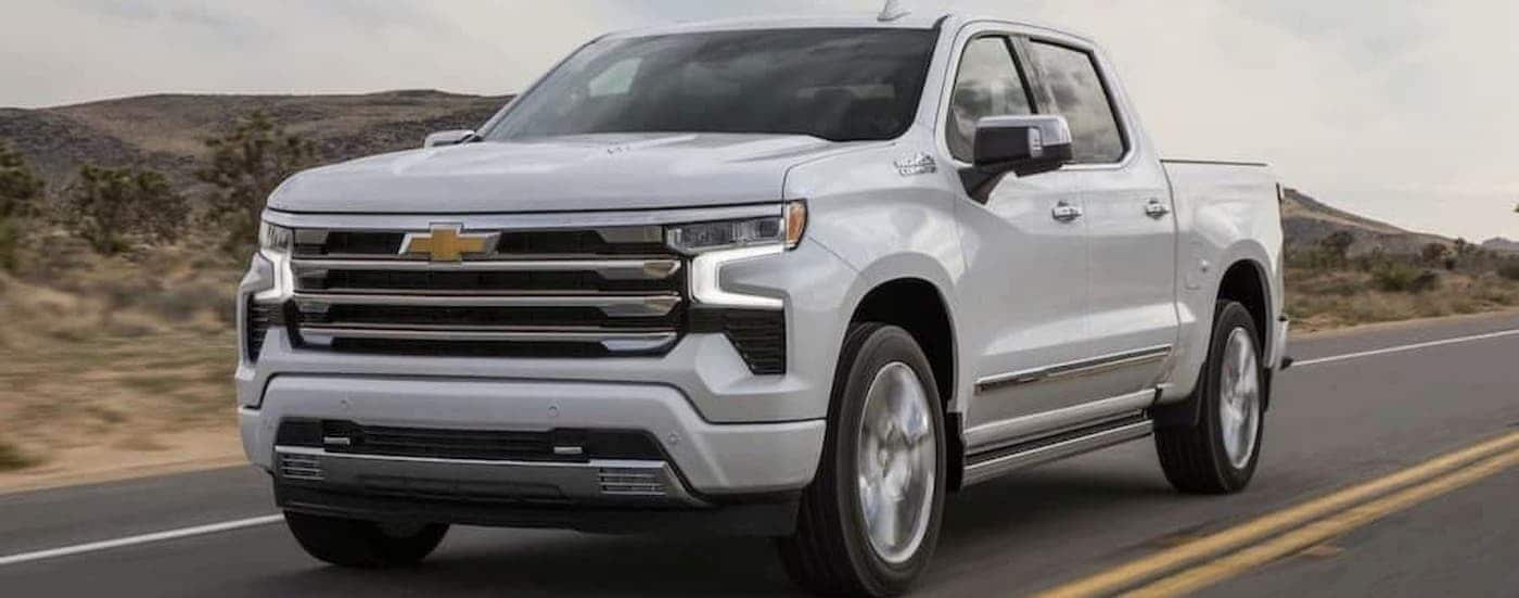 A white 2024 Chevy Silverado 1500 High Country is shown driving on a highway.