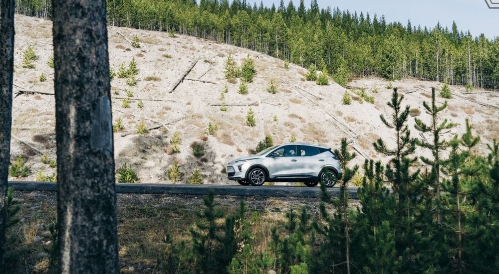 A silver 2023 Chevy Bolt EUV seen from a distance driving on a tree-lined road.