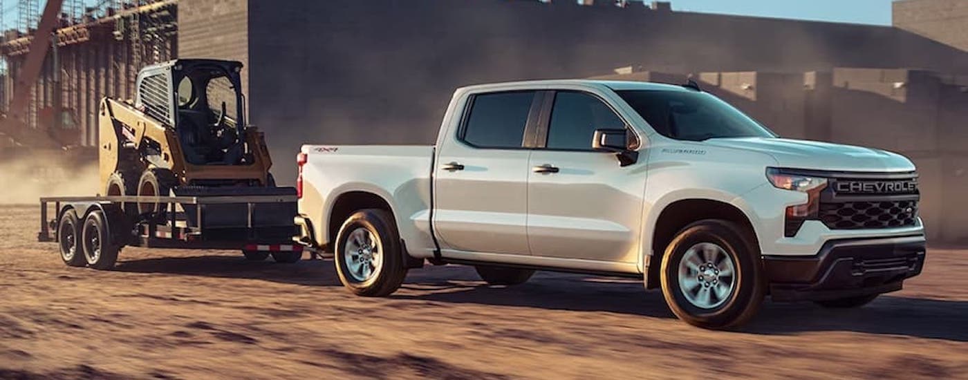 A white 2023 Chevy Silverad 1500 Custom is shown towing a skid-steer.