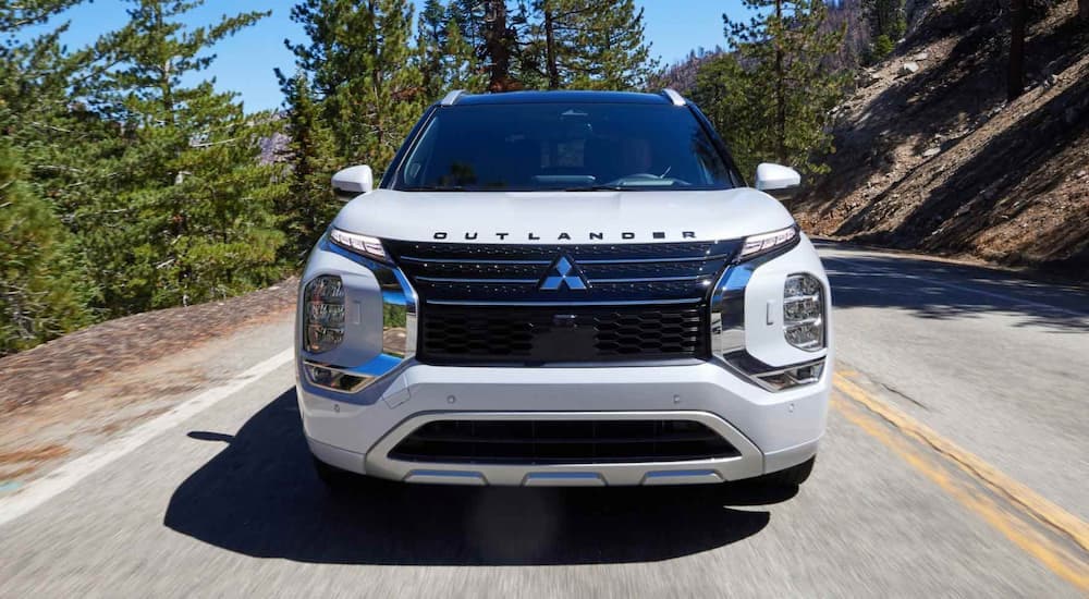 A white 2023 Mitsubishi Outlander PHEV is shown from the front driving on a tree-lined road.