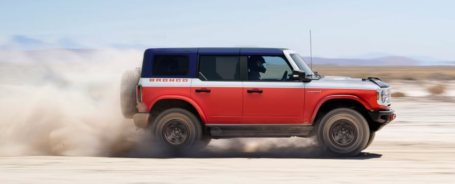 A red and white Ford Bronco in profile kicking up dust as it drives down a dirt road.