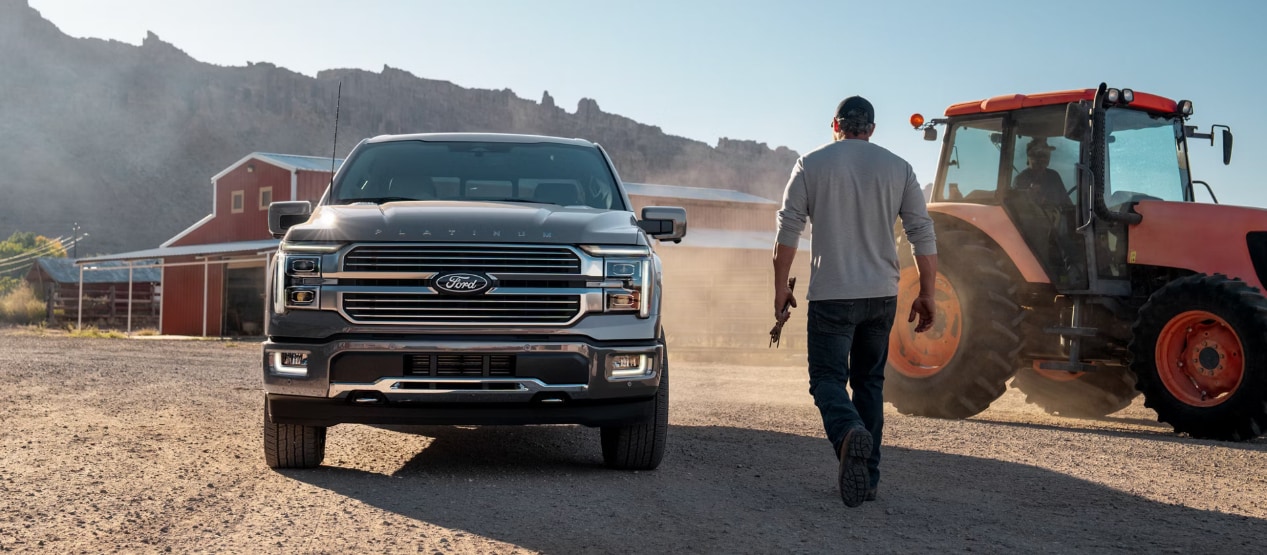 A front view of a gray Ford F-150 on a ranch by a red tractor and man walking towards it.