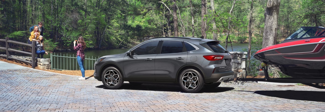 A gray Ford Escape towing a red boat while a woman in front of the car takes a photo next to a river on a brick trail.