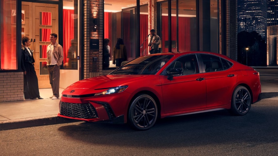 A red Toyota Camry parked by the curb outside a club at night.