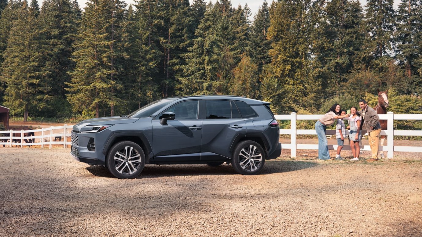 A blue Toyota RAV4 parked on a ranch near a family taking a photo with a brown horse.