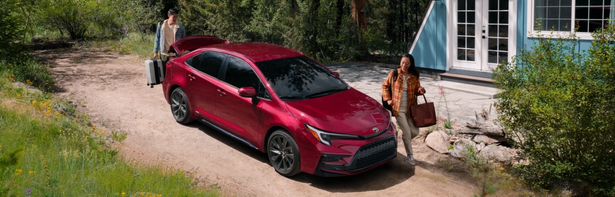 A red 2026 Toyota Corolla parked in front of a house with two people preparing to put their luggage inside.