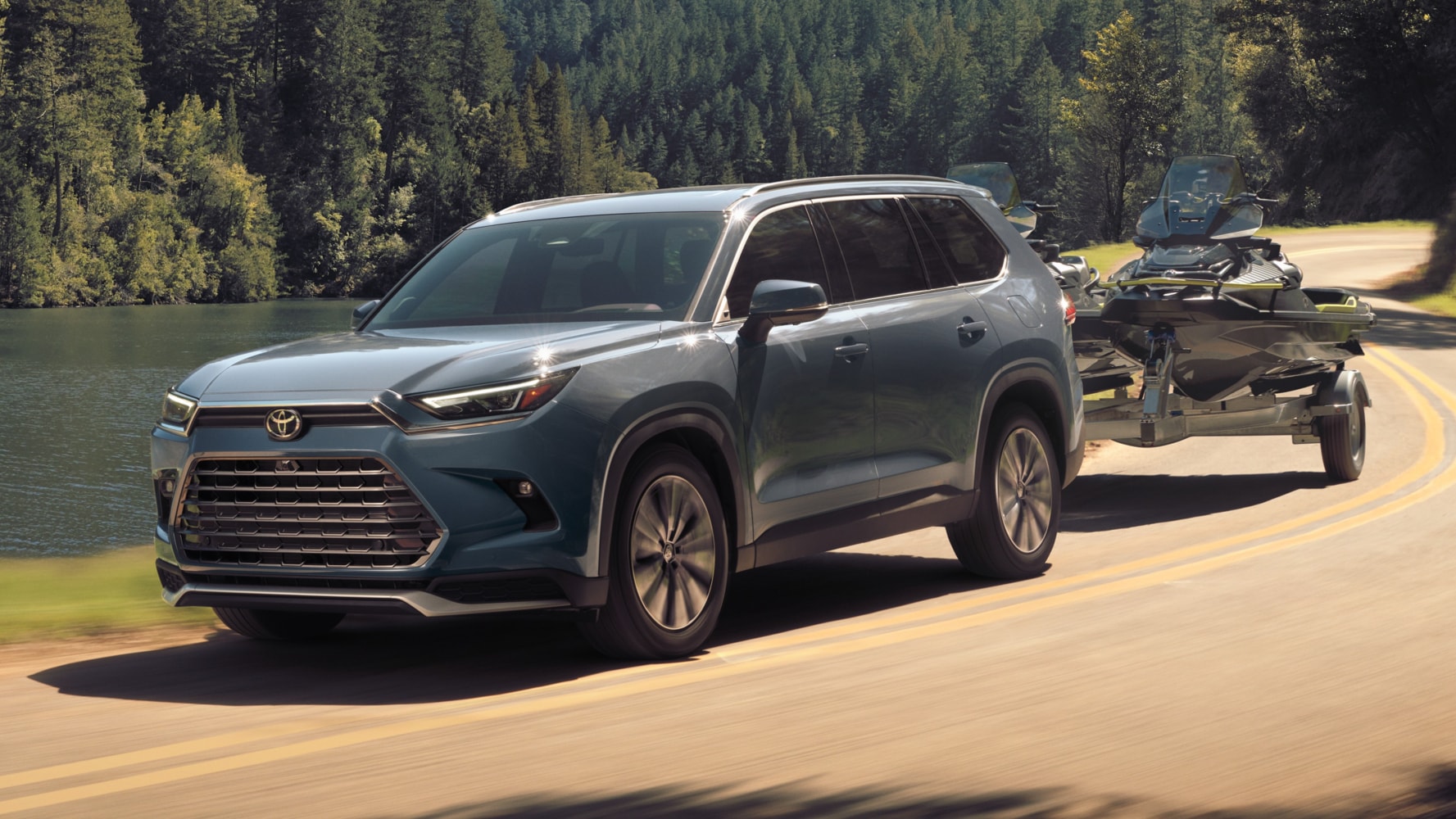 A light blue Toyota Grand Highlander driving down a road by a lake while towing a boat.
