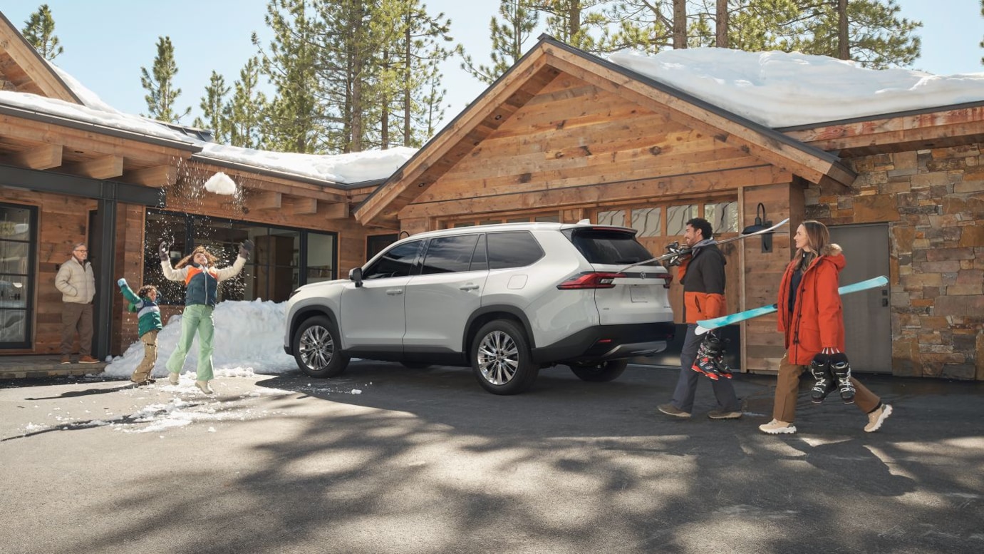 A gray Toyota Grand Highlander parked outside of a winter cabin as a family gathers around it.