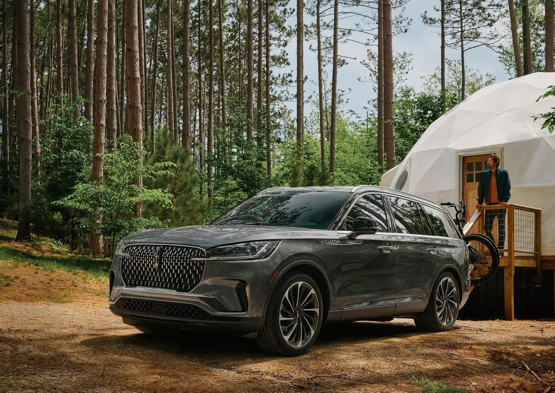 A grey Lincoln Aviator parked on a forest track beside a dome tent, a bicycle on its rack