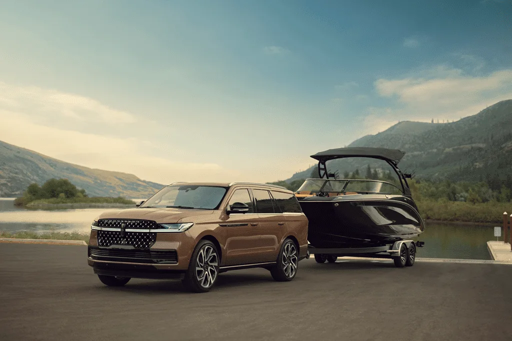 2026 Lincoln Navigator Black Label in a bronze finish towing a boat on a trailer beside a lake with mountains behind