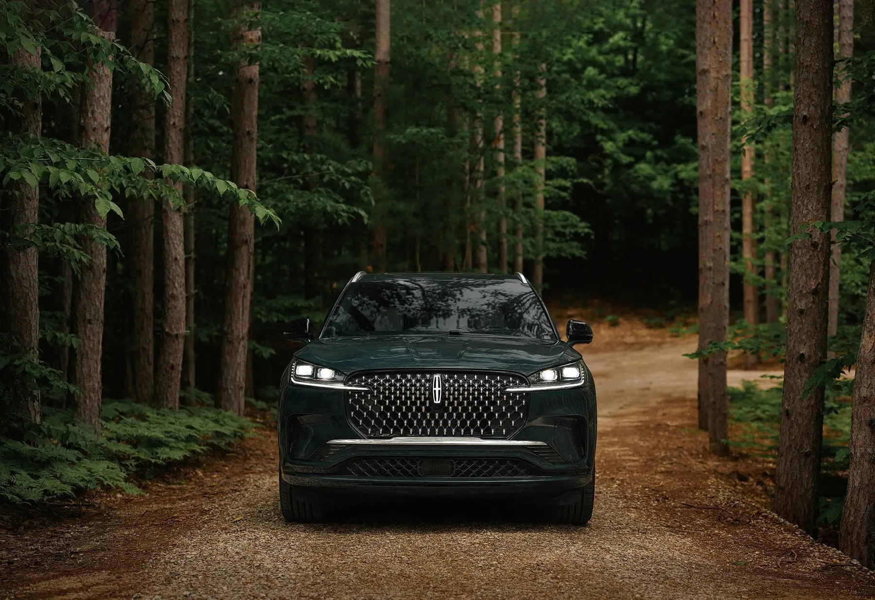 A dark green Lincoln Aviator head on with its headlights lit, on a gravel road through pine woods