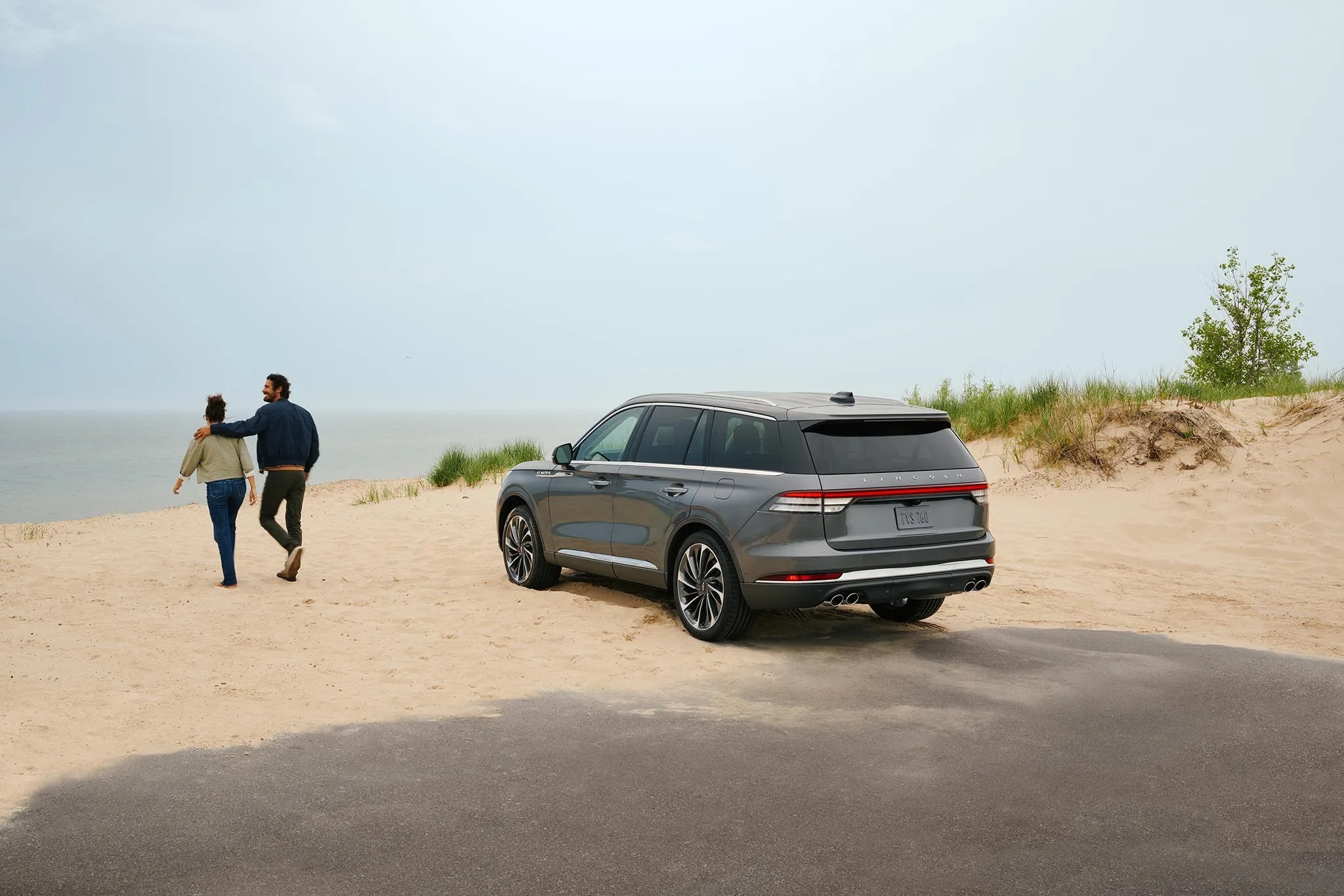 A gray Lincoln Aviator parked on the sand at the edge of a beach while a couple walk toward the water
