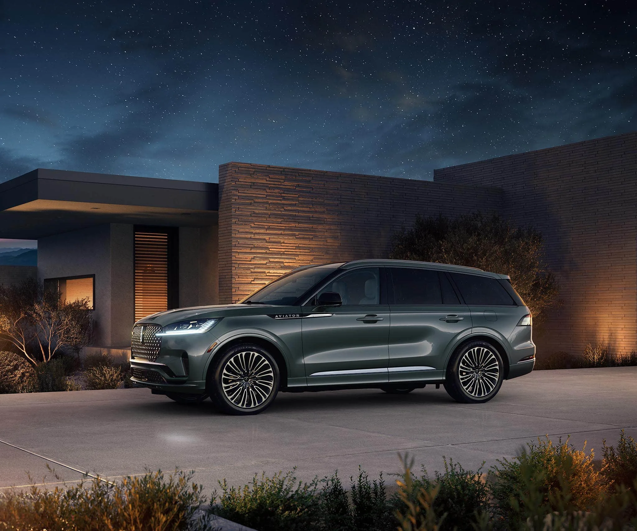 A dark green Lincoln Aviator Black Label parked on a driveway outside a modern brick house under a starry night sky