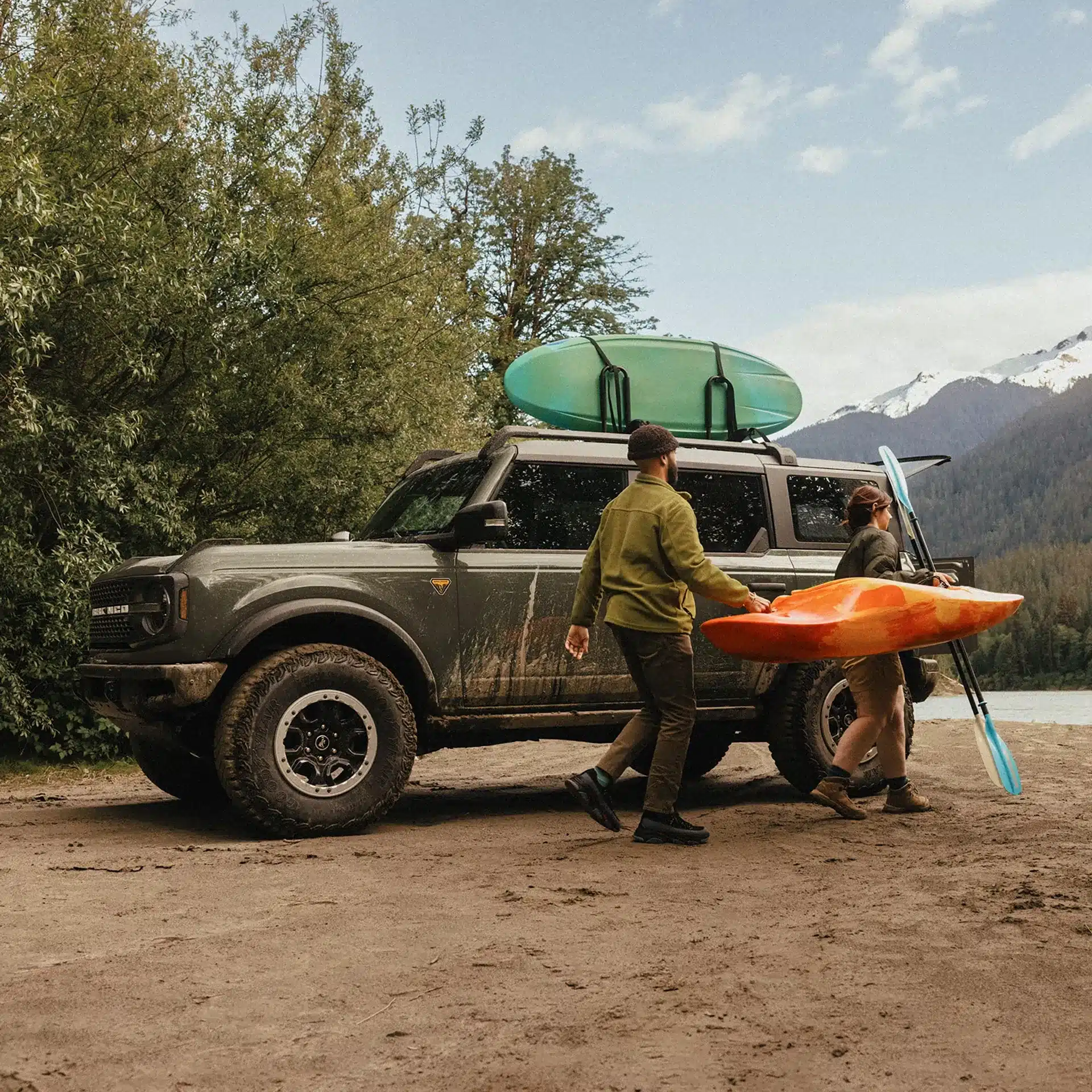 A muddy, dark green 2026 Ford Bronco parked on a dirt bank near a lake and mountains. A man and a woman walk past the driver's side, carrying a bright orange kayak and a paddle. A green paddleboard is secured to the roof rack of the SUV.