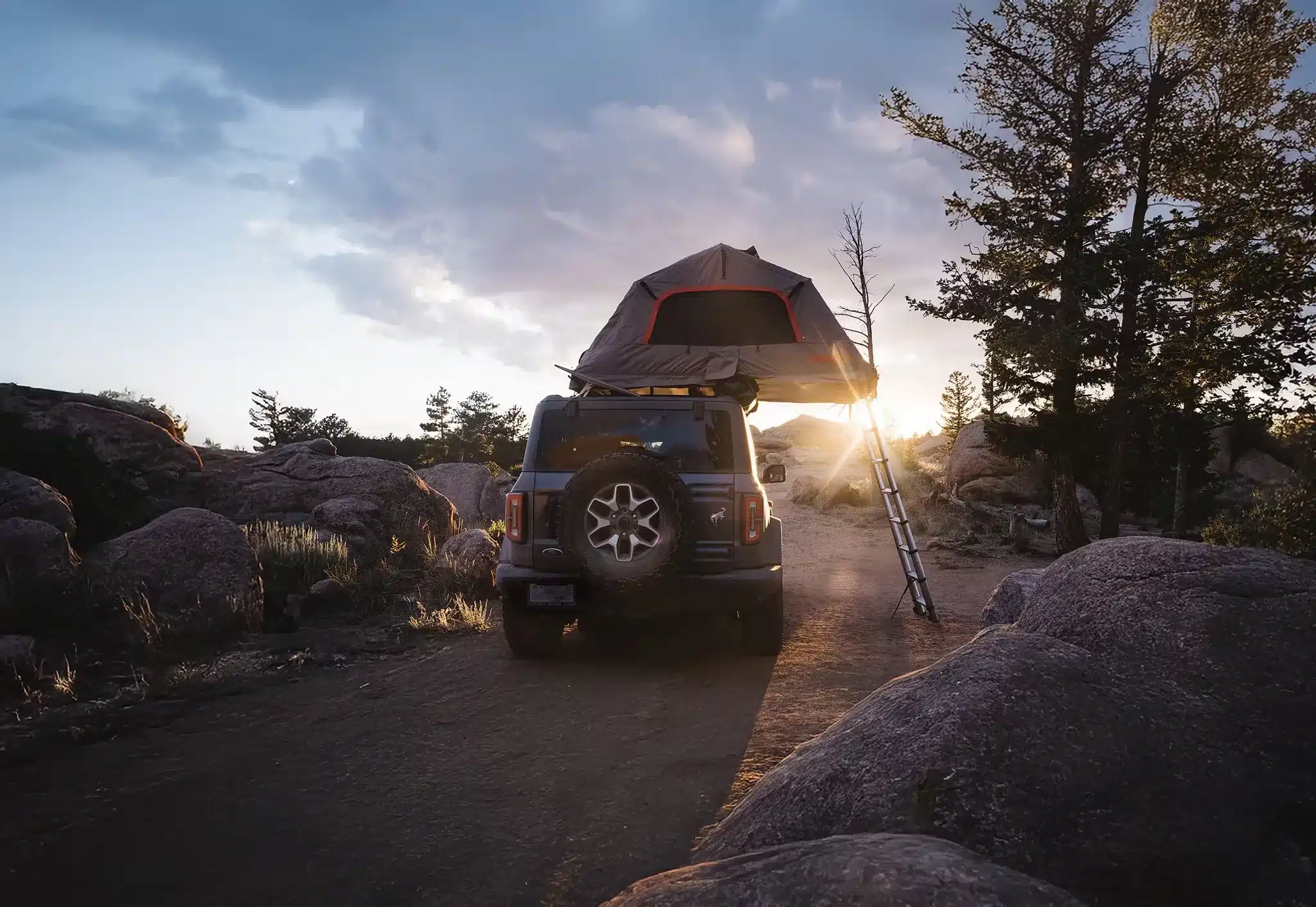Rear view of a dark 2026 Ford Bronco parked on a dirt road at sunset, with a rooftop tent deployed on the roof rack and a ladder leaning against the rear tire.